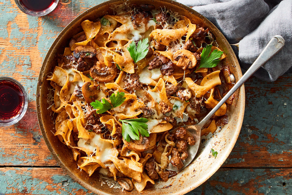 A bowl of French Onion Beef and Noodles topped with melted Gruyère cheese, garnished with fresh thyme, and served with a slice of crusty bread